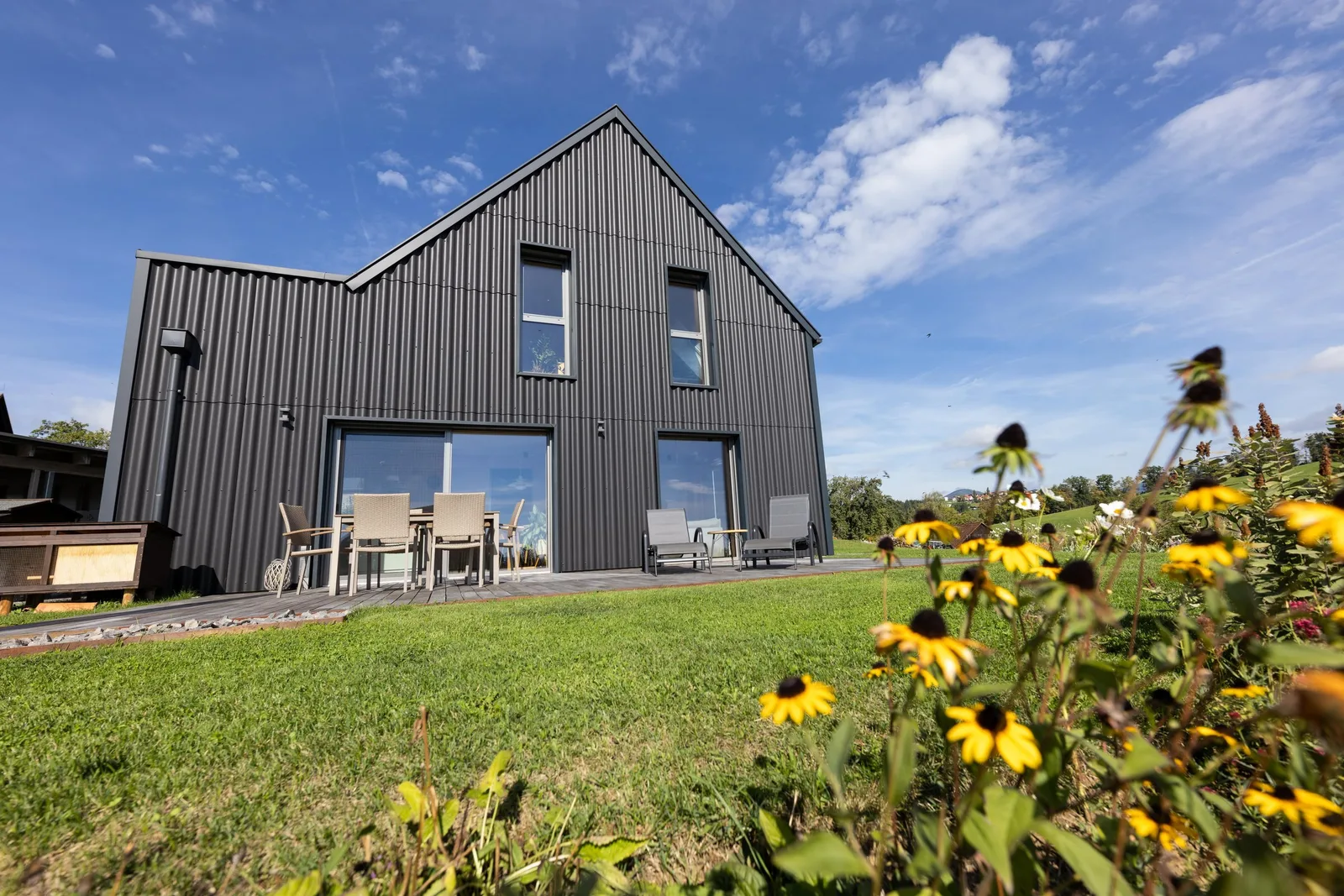Dunkles Wohnhaus mit gewelltem Dach und gewellter Fassade, gelbe Blumen im Vordergrund