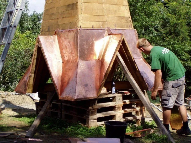 Mitarbeiter in grünem Shirt an einem Turmhelm mit Kupferverkleidung, der auf Paletten aufgebaut ist