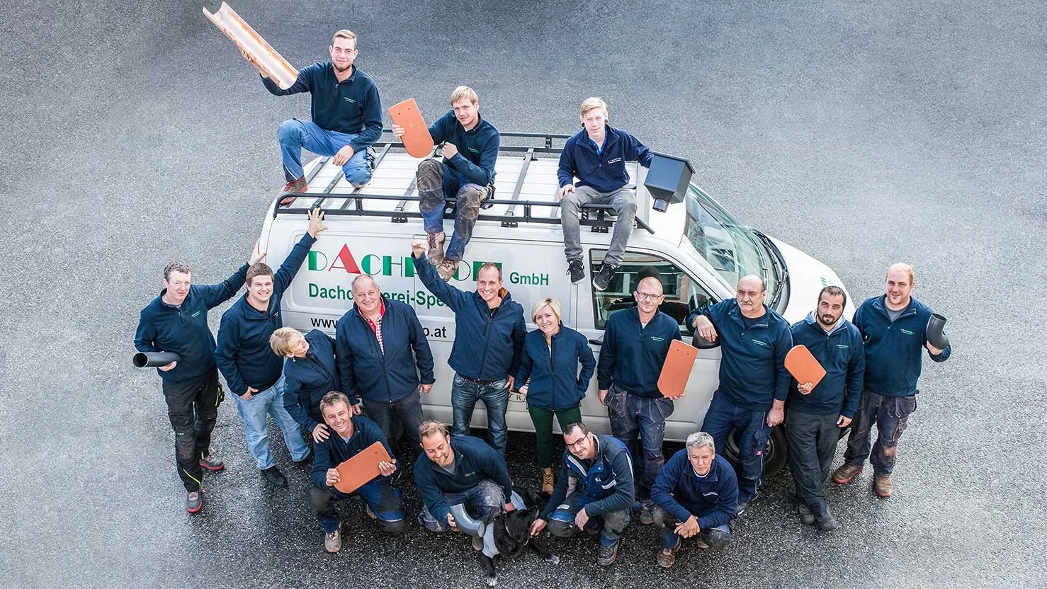 Das Dachprofi-Team um den Firmenbus, einige sitzen auf dem Dach des Busses, mit Dachziegeln und Rinnenstücken in den Händen